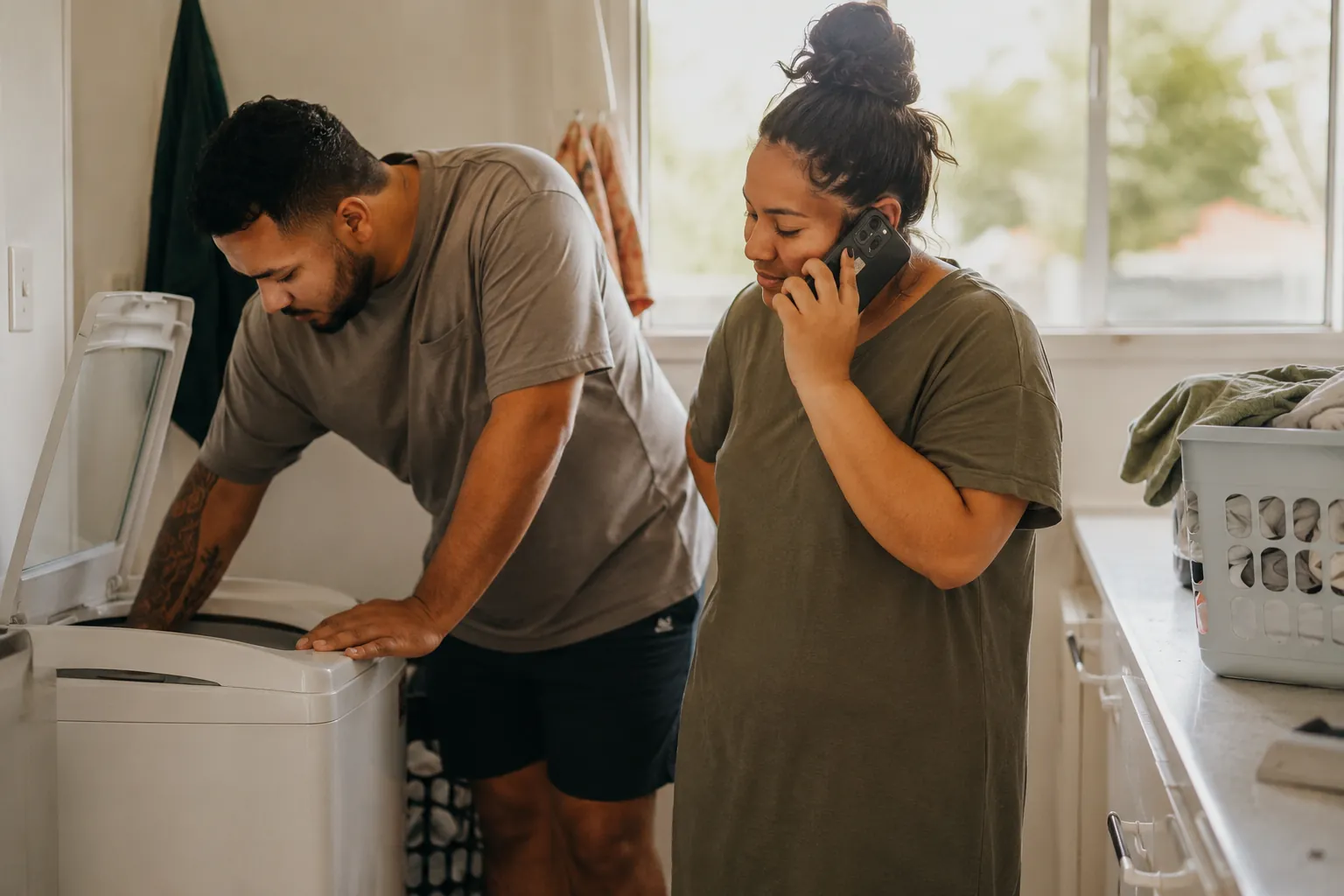 A couple in their laundry, one leaning over an open top-loader washing machine while the other makes a phone call