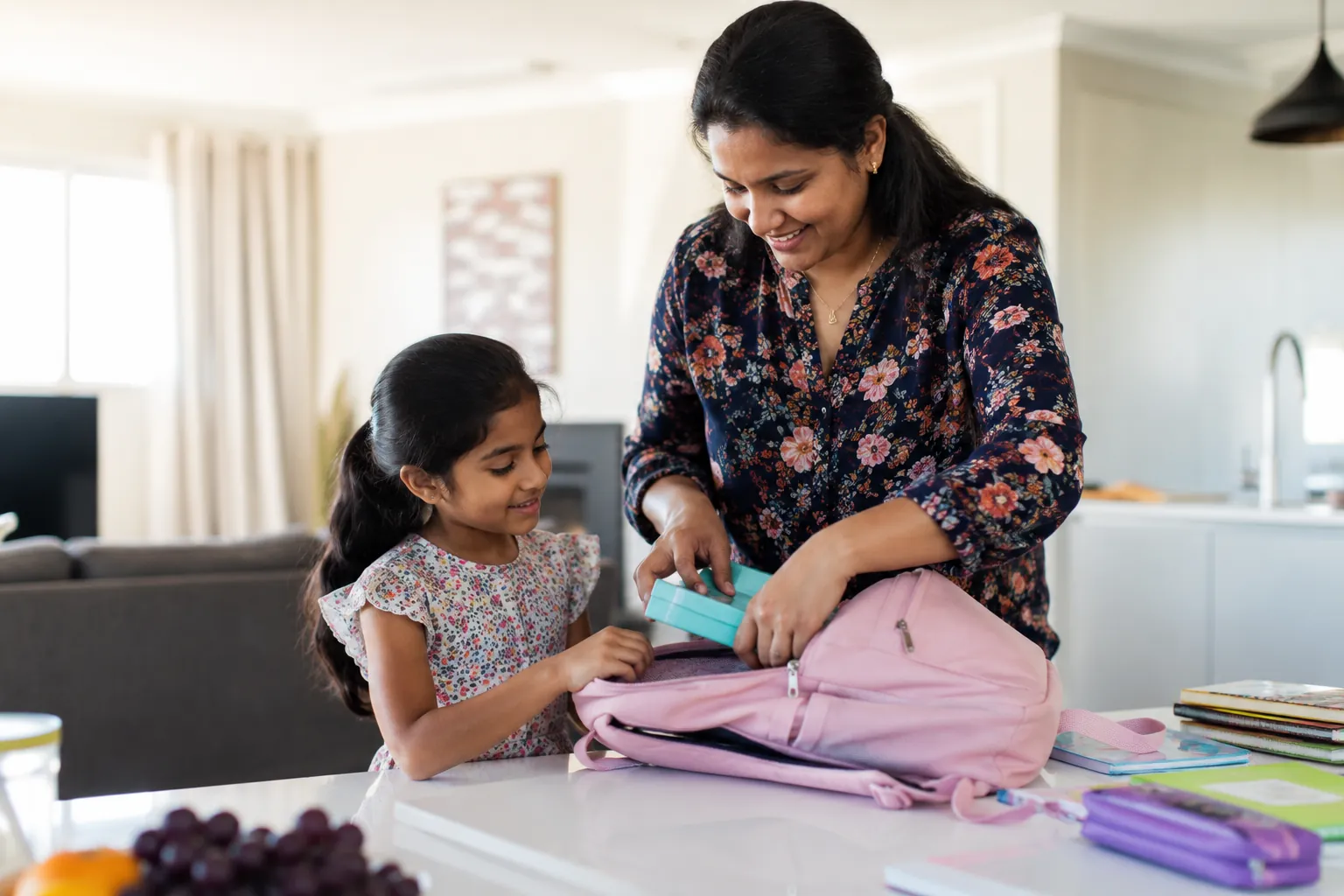 A mother and her daughter pack a lunchbox into a pink school backpack at the kitchen bench